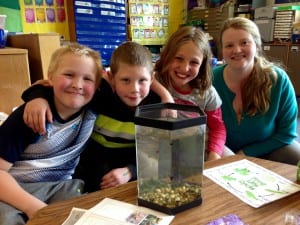 Eric Thynes, James Clark, and Iris Case sit with their reading teacher, Lisa Schramek near the tadpole tank. Photo/Angela Denning