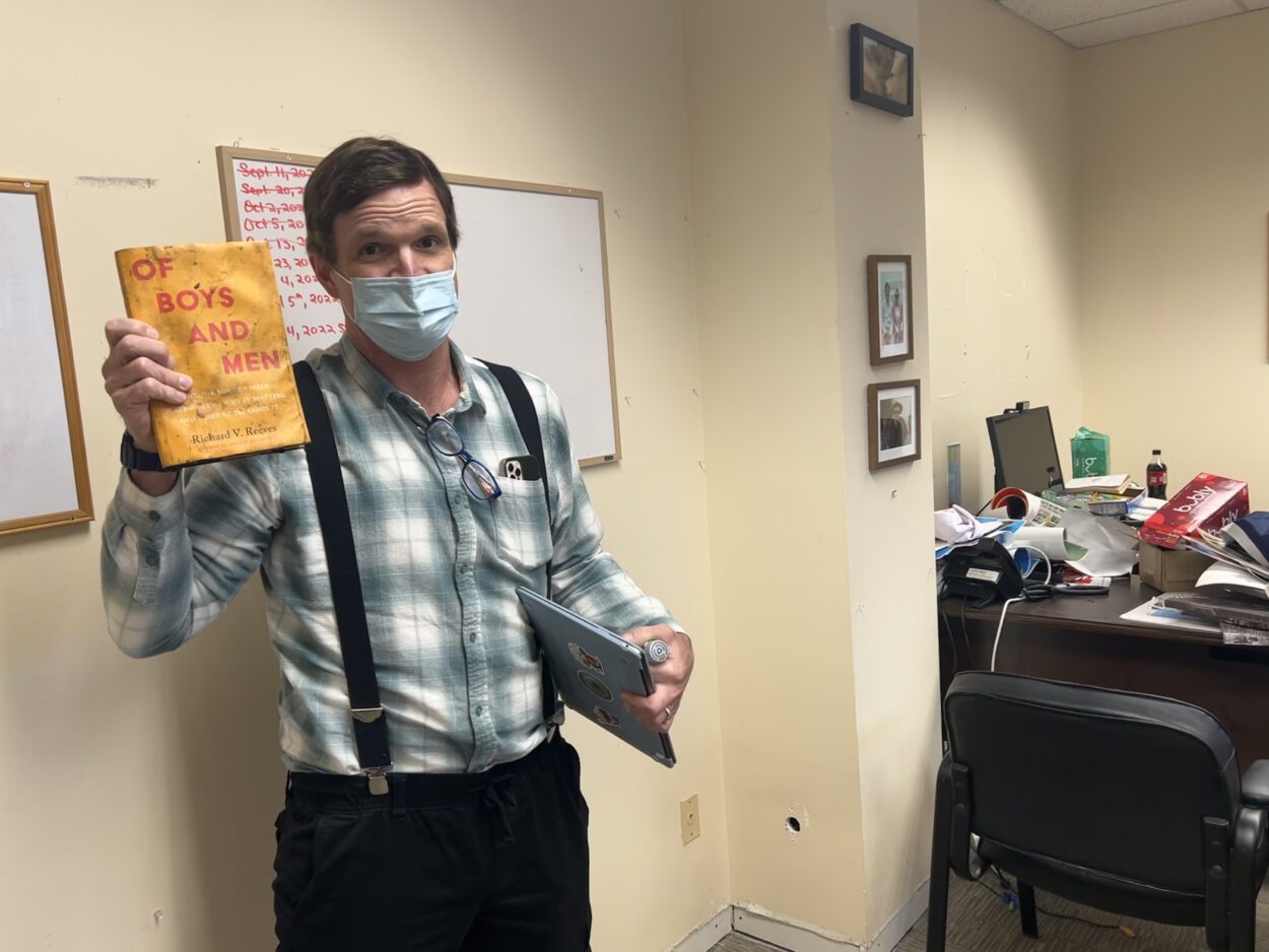 Ambler Moss stands in his office. He is holding up a book titled "Of Boys and Men," which appears to have sustained severe water damage.