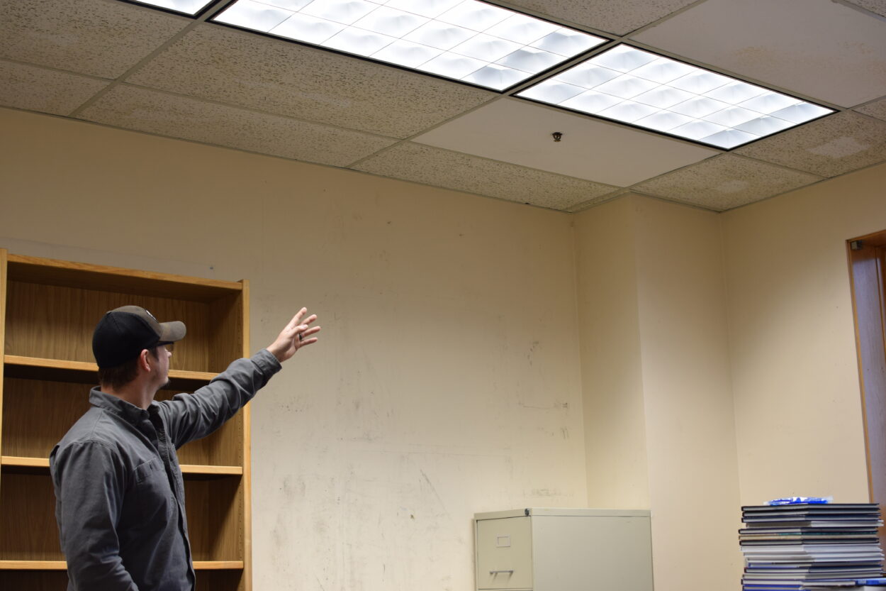 Aaron Buller gestures toward a sprinkler head in a school office. The office is empty, save for a stack of textbooks, a filing cabinet, and a bare shelf.