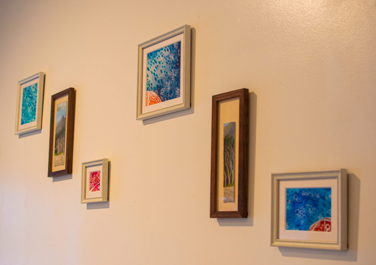 Six small framed paintings hang in two parallel diagonal lines. The paintings in the first and third rows are of abstract seascapes and the middle two are long, narrow paintings of forested landscapes.
