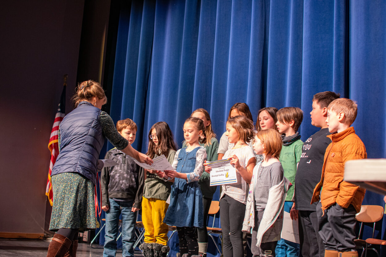 Jill Norheim and twelve elementary students are gathered on a stage. Norheim hands a young girl a certificate.