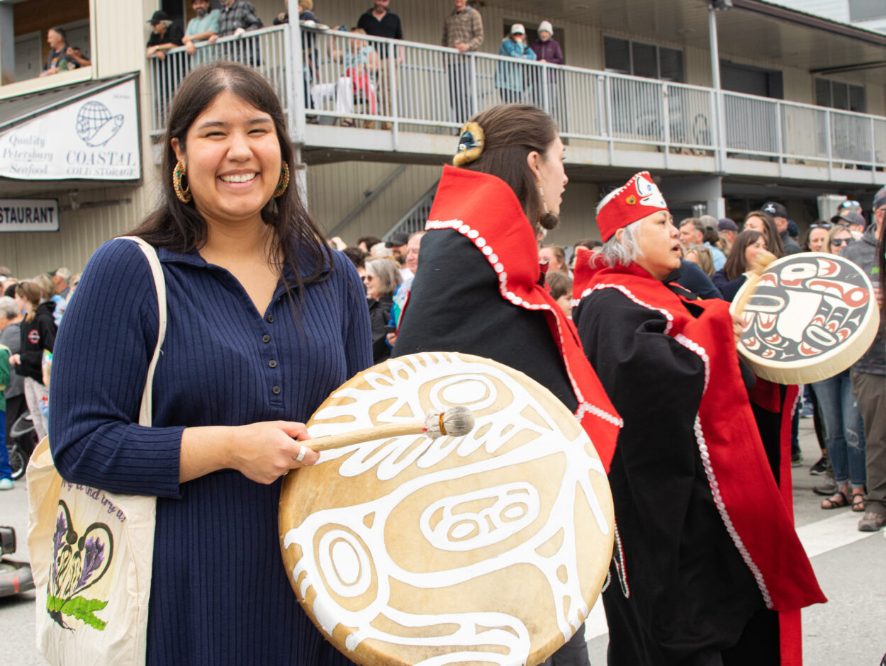 Avery Herman-Sakamoto smiles as she walks with a leather drum in her hand. She is flanked by two Southeast Alaska Indigenous dancers wearing traditional capes.