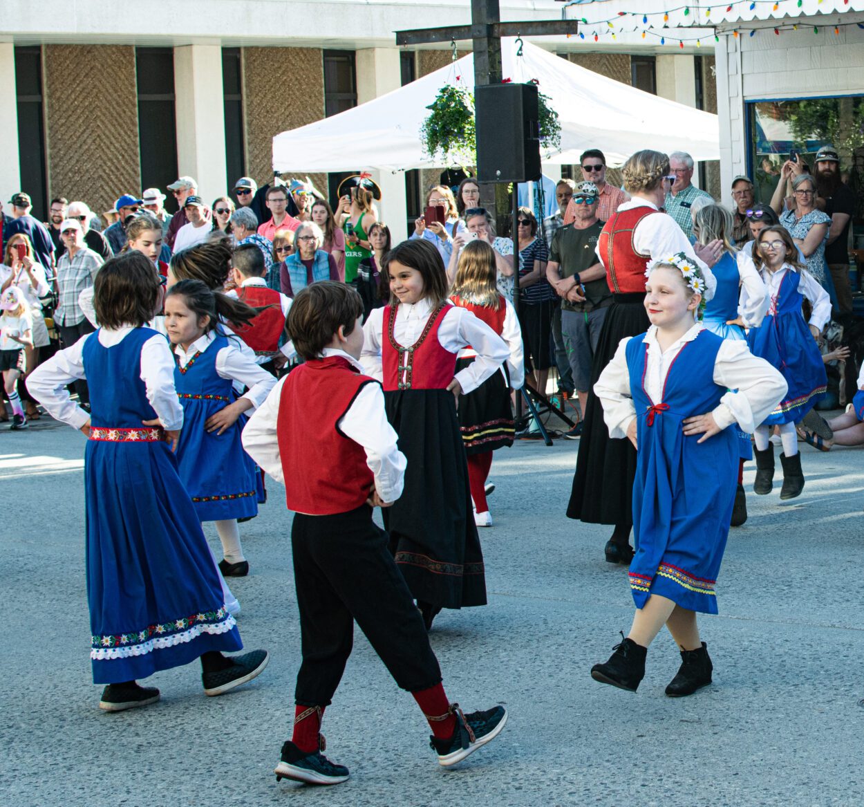 Three girls and one boy wearing Norwegian folk costumes dance with each other in a crowded street.
