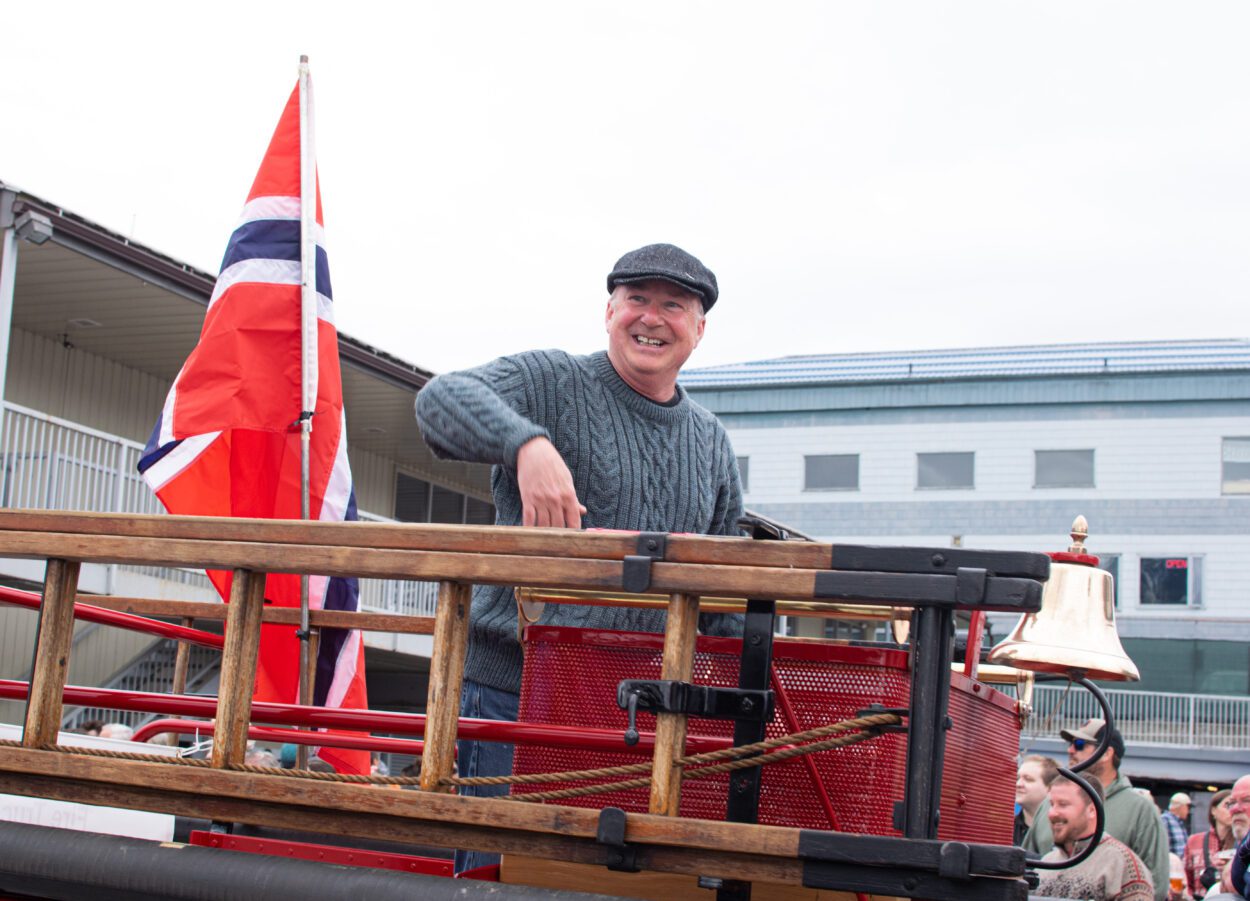 Tom Abbott stands on top of a fire truck, smiling. There is a Norwegian flag behind him.