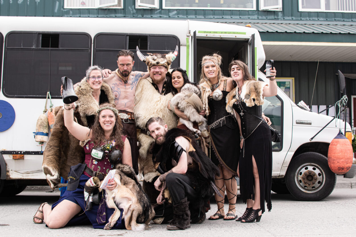 A group of vikings and valkyries dressed in furs stand in front of a small bus.