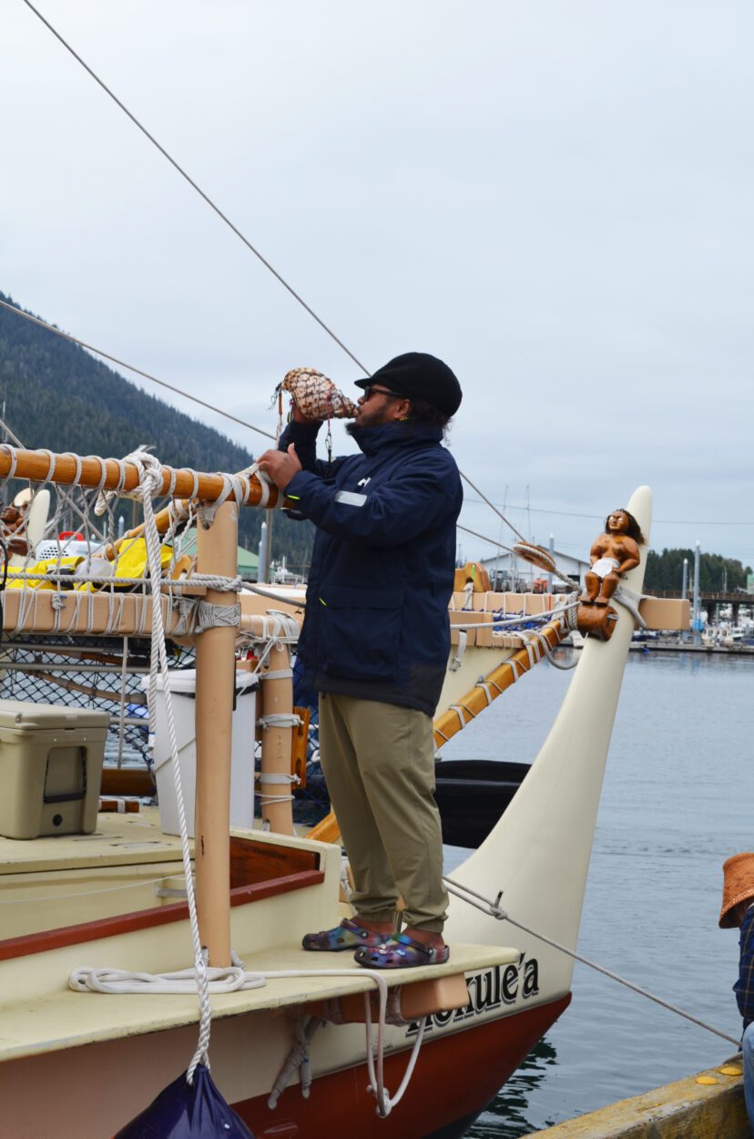 A Hōkūleʻa crew member holds a decorated conch shell horn to his lips. He is standing aboard the Hōkūleʻa at Petersburg's South Harbor.