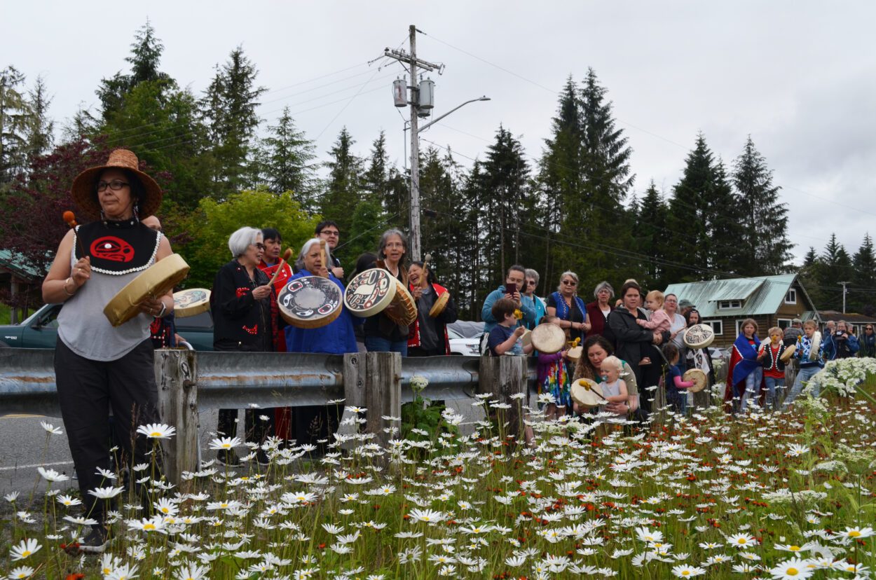 A couple dozen people lined North Nordic Drive on June 25th. Some are dressed in Lingít regalia and are playing hand drums. They are leaning against a roadside guardrail and there is a large patch of daisies in front of them.