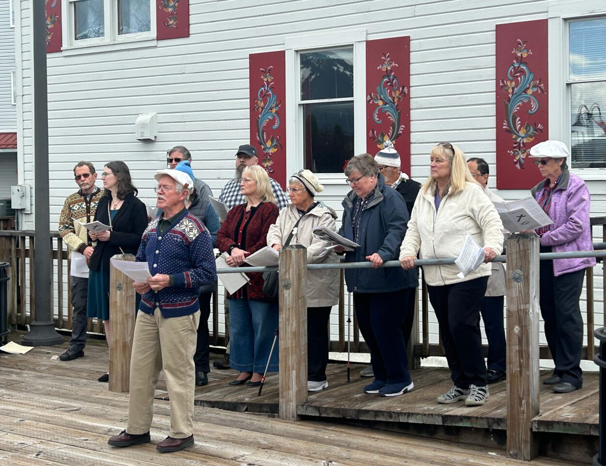 Grant Trask stands in front of a twelve-person choir on the boardwalk adjacent to the Sons of Norway Hall. They all hold sheets of music in their hands. Trask is facing away from the singers and speaking to the crowd.