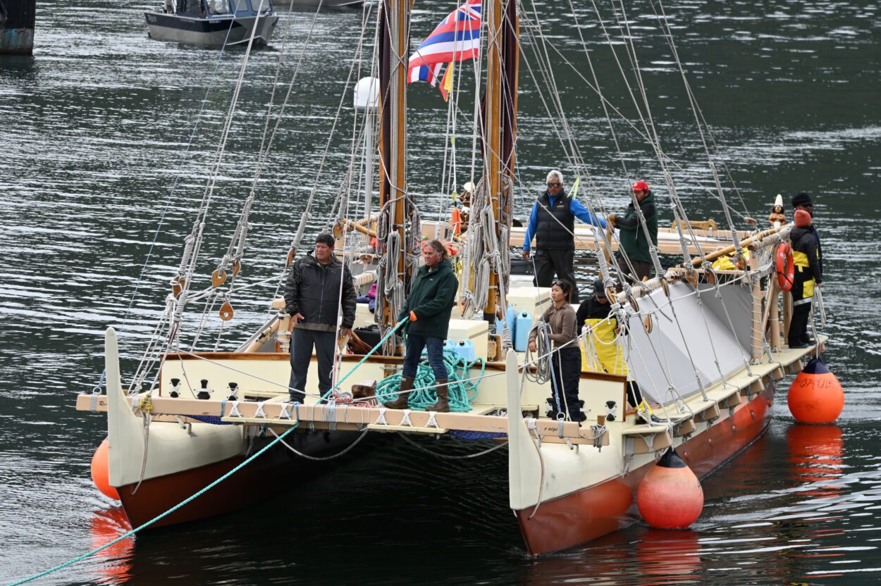 The bow of the Hokulea points at the camera. Ten crew members are in view, gathering ropes and preparing the canoe to dock in Petersburg's South Harbor. Mark Ellis, the canoe's captain, stands at the very front.