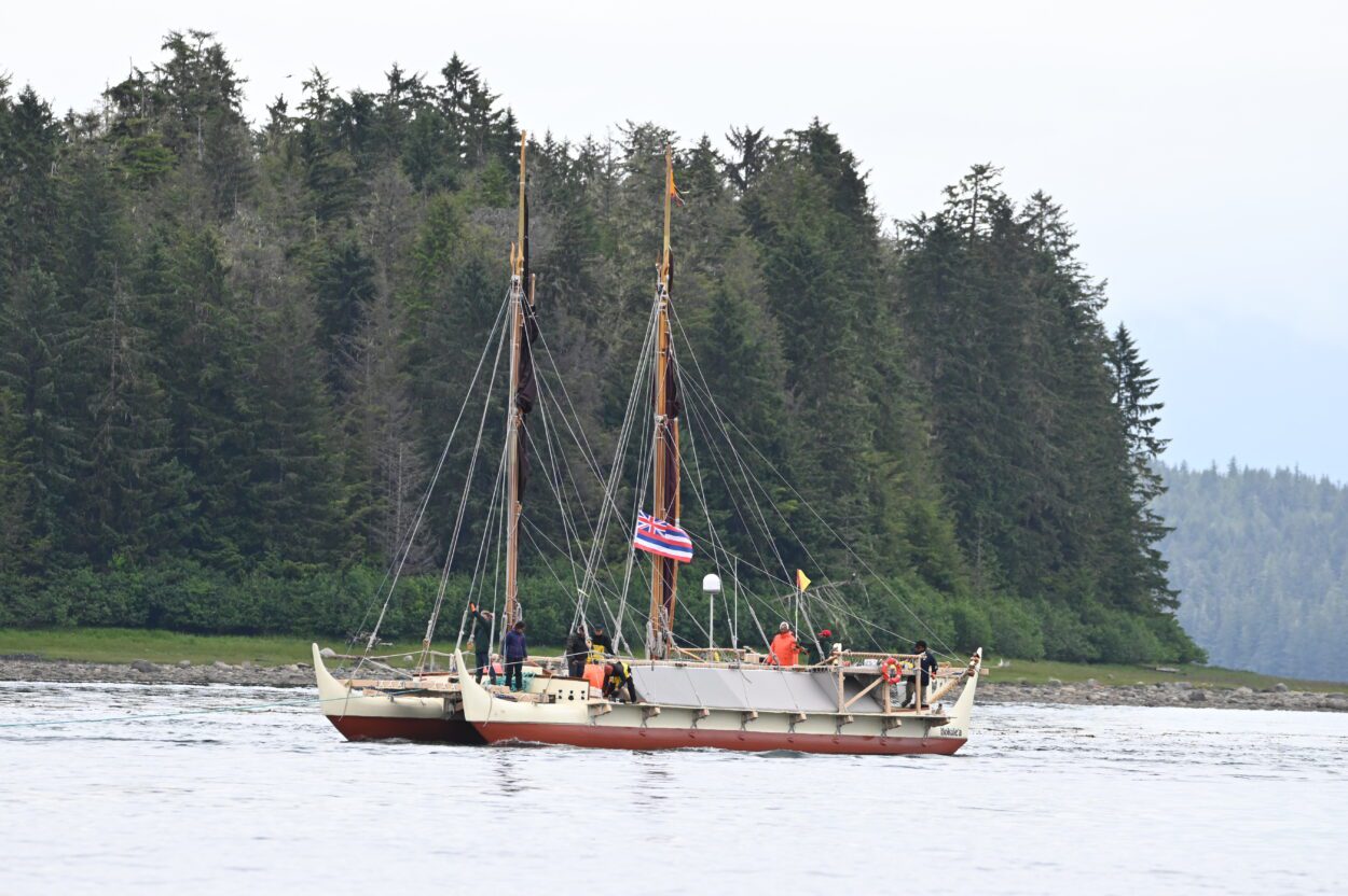 The Hōkūleʻa, a twin-hulled canoe, sailing through the Wrangell Narrows. There are about eight crew members in view on board and an evergreen forest on the coastline behind the canoe. The state flag of Hawaii is raised halfway up one of the two masts.