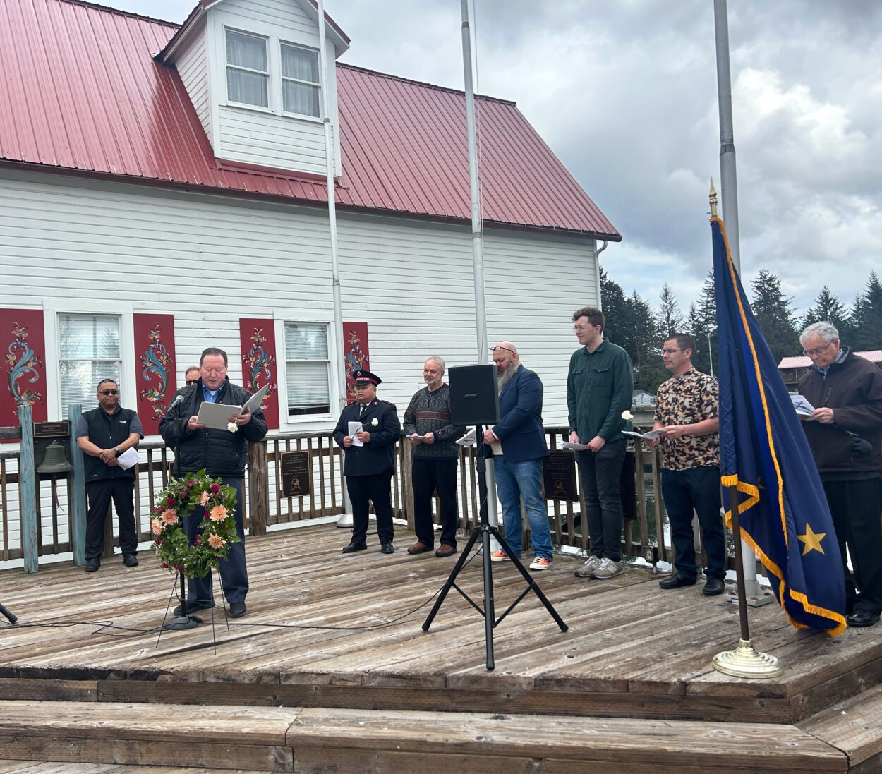 Nine religious leaders of the Petersburg Ministerial Association stand on the boardwalk adjacent to the Sons of Norway Hall. Their heads are bowed in prayer. Pastor Pastor Lee Corrao stands at the edge of the platform in front of a microphone. There is a wreath of flowers propped up by his knees.