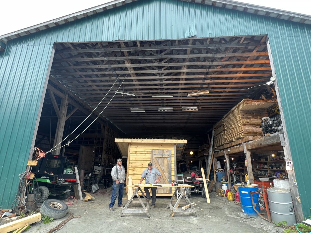 Brett Martin and Mike Duman stand at the threshold of a large warehouse. Brett Martin is leaning on a workbench and Mike Duman stands to his right. Behind them, there is a small wooden shed.