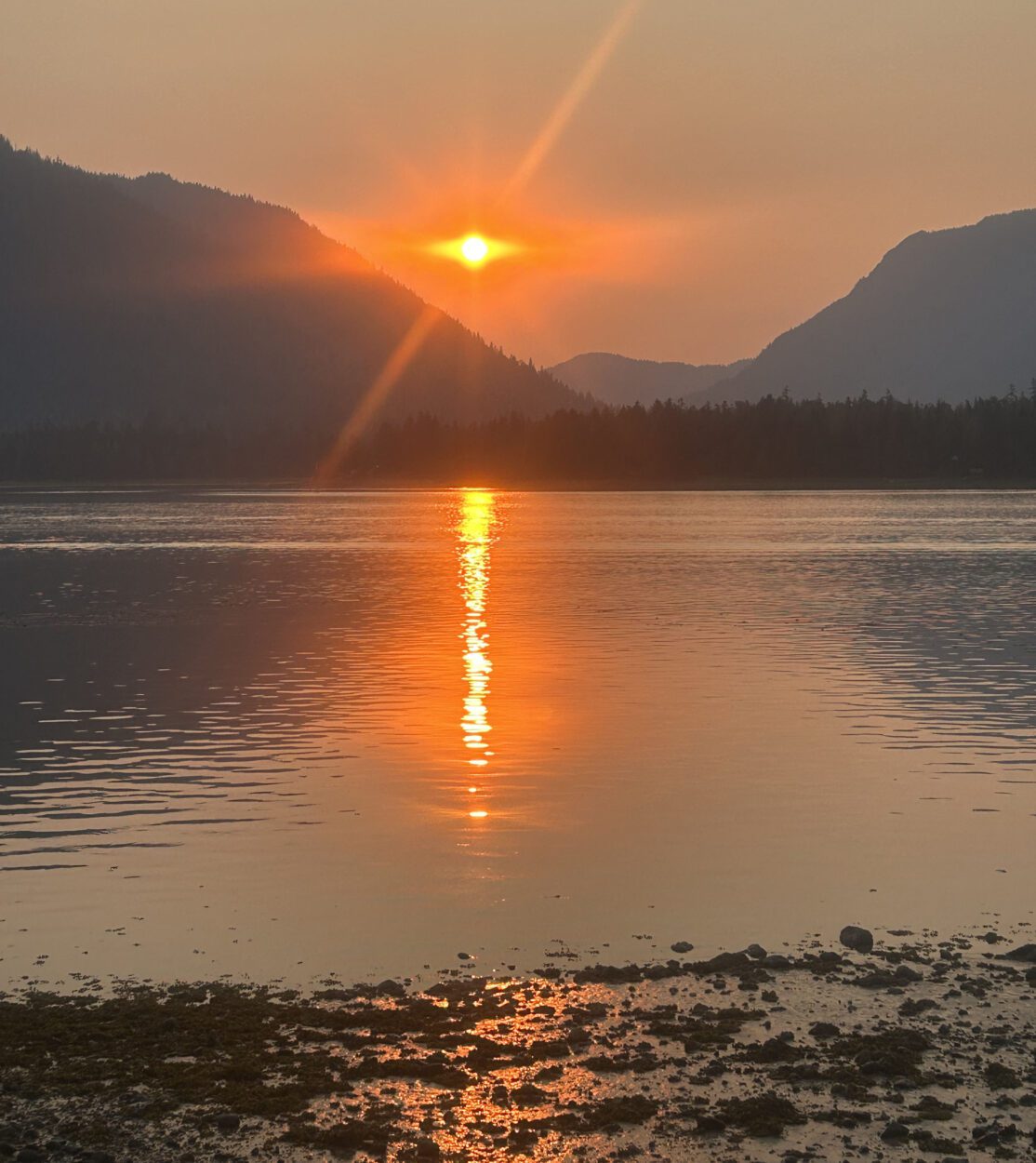 The sun setting between two large mountains behind a channel of water. The shoreline is visible in the foreground. There is a slight haze in the sky that turns the setting sun bright orange.