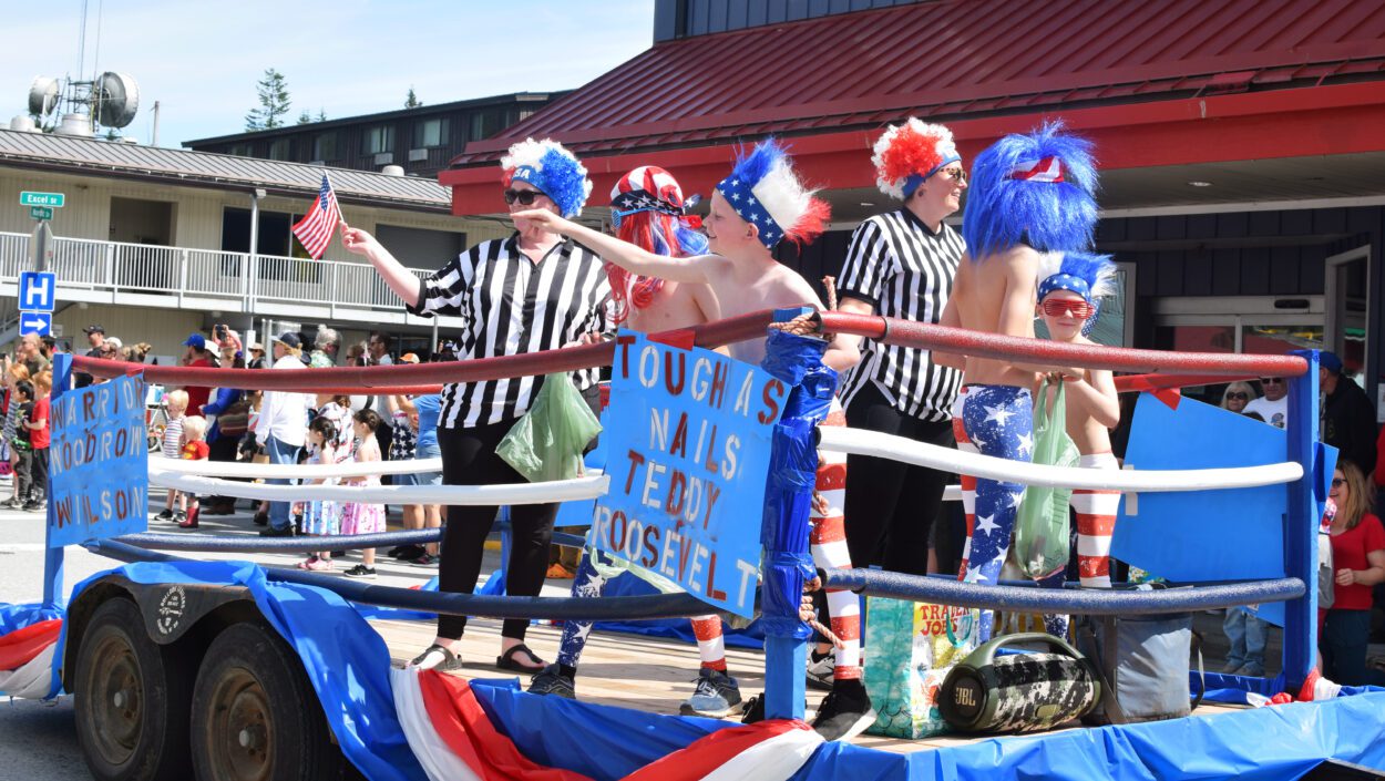 A Fourth of July Parade float designed to look like a professional wrestling ring. In the ring, there are three children dressed as America-themes professional wrestlers and three adults dressed as referees. The float is passing down Petersburg's Main Street, just past Excel Street.