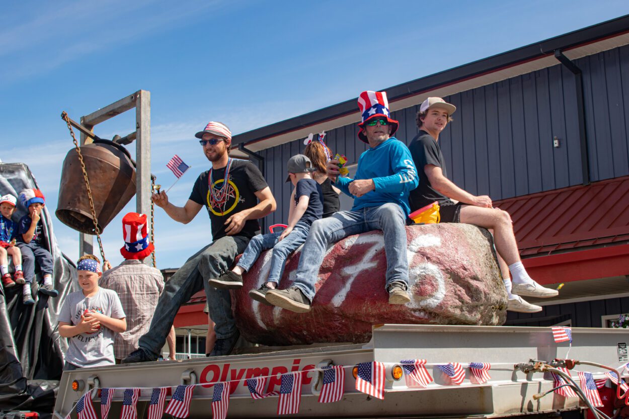 A group of nine adults and children sit on a parade float pulled atop the flatbed of a semi truck. Four of them sit on a large boulder, spray-painted with the date: 1776. A man off to one side rings a giant metal bell.