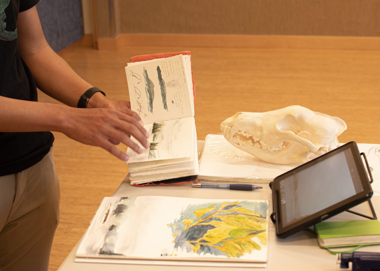 Trenton Jung's hands are pictured holding a handmade sketchbook, in which he has painted some landscapes. He is resting the sketchbook on an end table. There are art supplies, a wolf skull, and an electronic tablet on the table's surface.