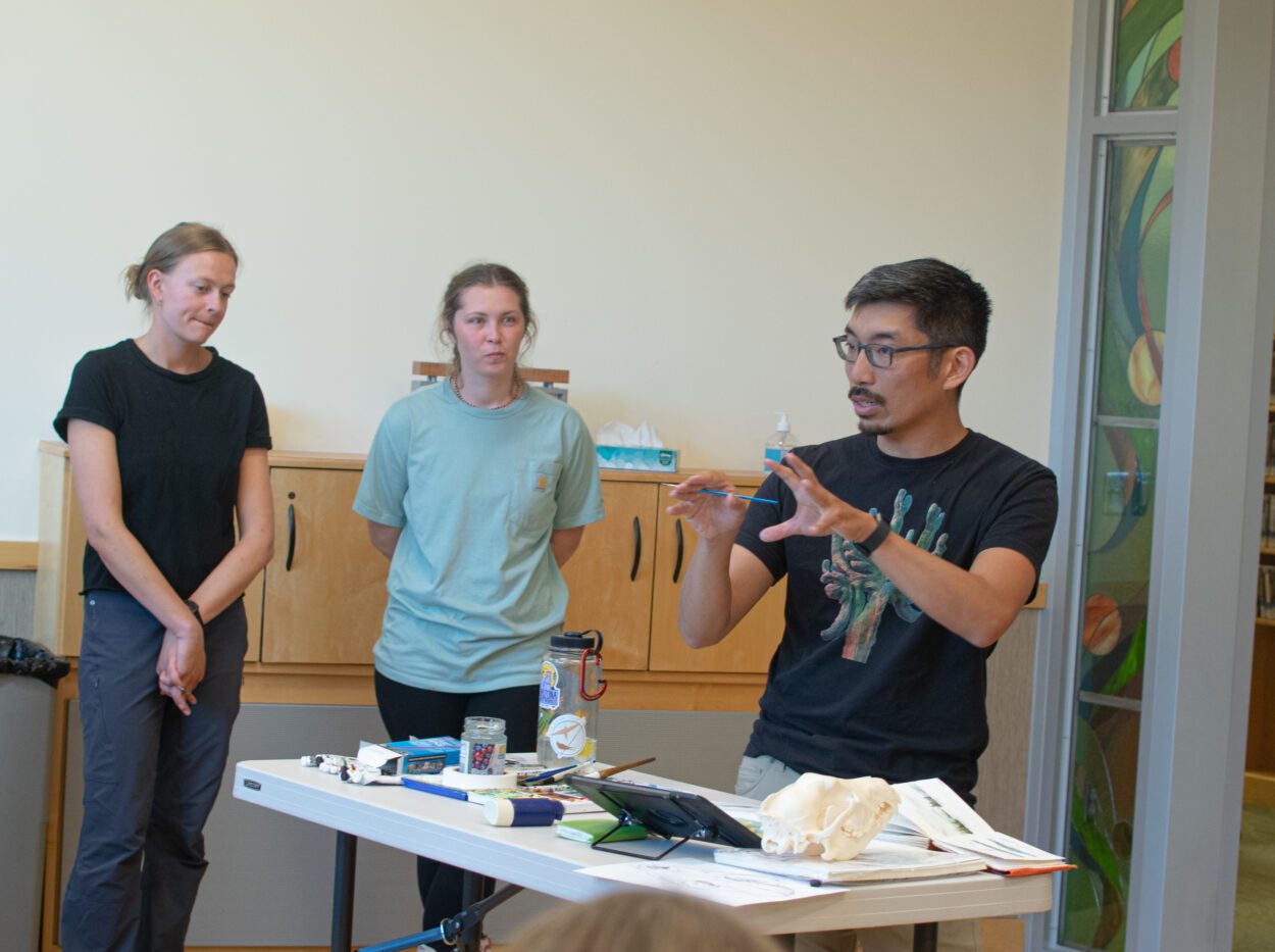 Trenton Jung is standing in front of an end table full of art supplies, explaining something to his class. Two women stand off to the side.