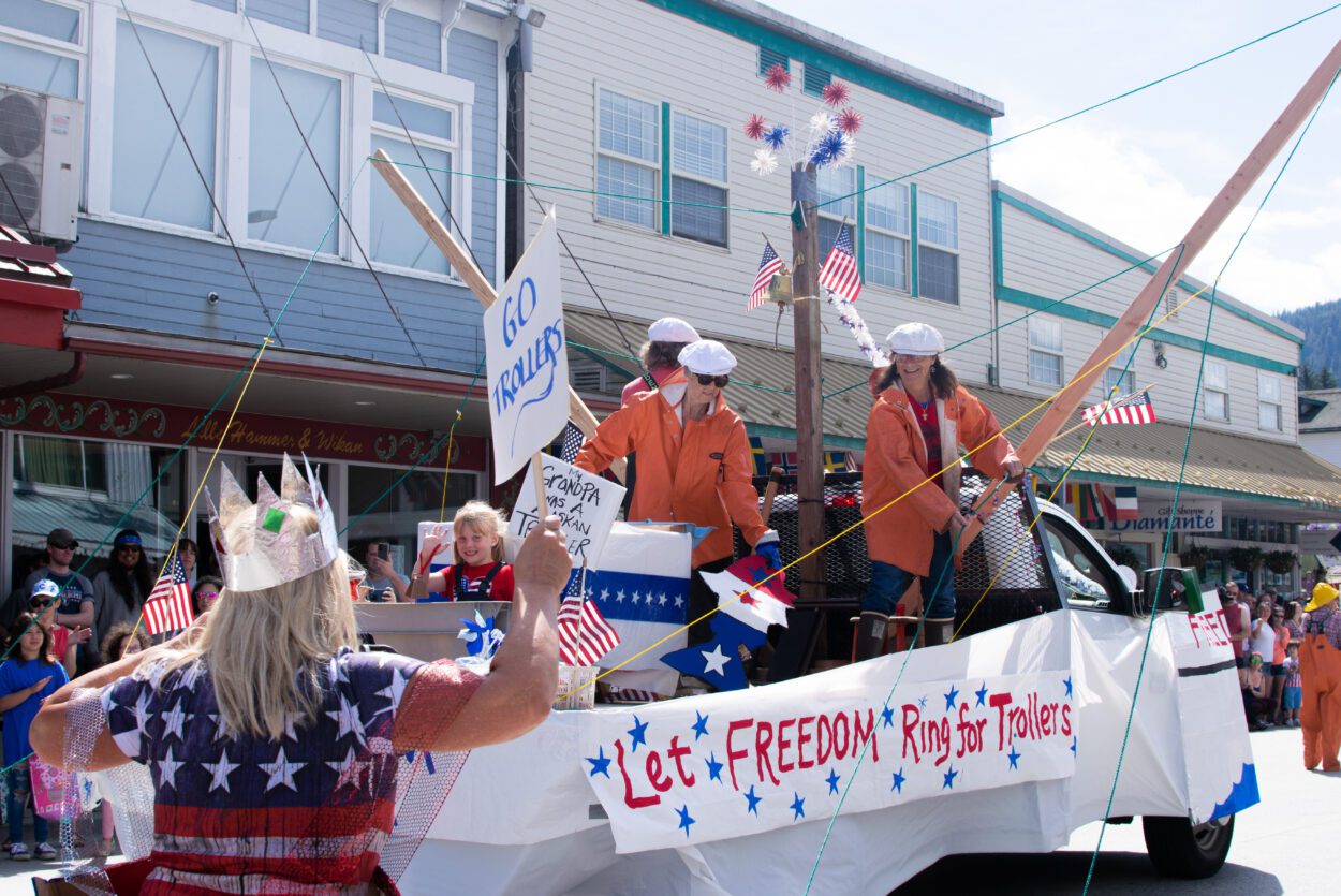 A parade float made to look like a trolling vessel. Three women dressed in orange fisherman's coats cast lines from atop the boat.