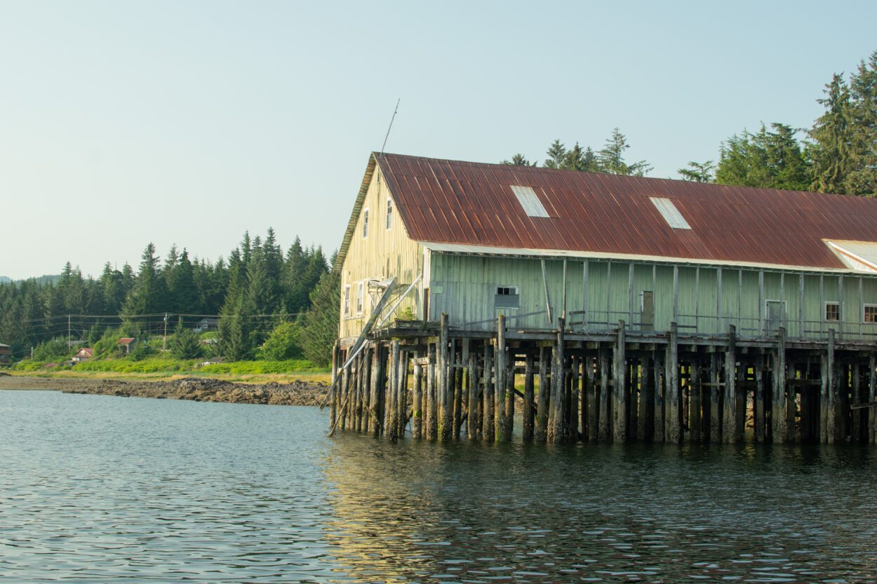 A shoreline with a building on stilts jutting into the water. There is a dense forest in the background.